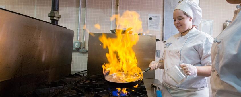 A cooking student dressed in her chef uniform at a stove range with flames ascending from a skillet in her hand.